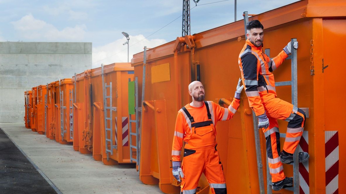 Two refuse collectors in orange high-visibility clothing are standing at disposal containers.